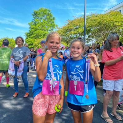 Two girls in blue Girls on the Run shirts embrace with smiles and 5k finisher's medals